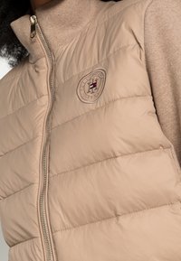 Beige lichtgewicht gewatteerde vest met gequiltde textuur, hoge kraag en metalen rits; heeft geborduurd logo op de linkerborst.