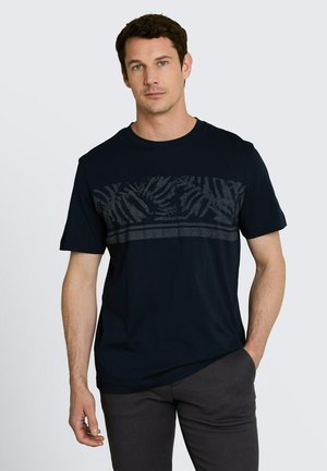 Man draagt een marineblauw T-shirt met korte mouwen en een donker bladpatroon over de borst, gecombineerd met donkergrijze broek, staand tegen een effen achtergrond.