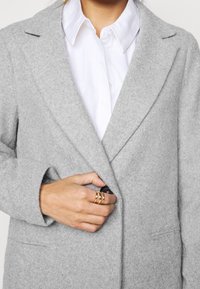 Henkilö kiinnittää napin vaaleanharmaasta villatakkista valkoisen kauluspuseron päältä, kädessä on kolme kultasormusta oikean etusormen ympärillä.