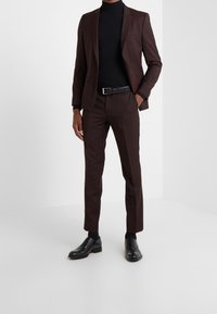 Completo marrone con una vestibilità aderente, dolcevita nero strutturato, cintura nera e scarpe nere lucide. I pantaloni hanno una vestibilità slim e gamba dritta.