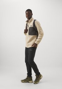 Beige fleece takki harmaalla rintataskulla, mustat housut ja vihreät vaelluskengät. Takissa on vetoketjullinen etuosuus ja korkea kaulus.