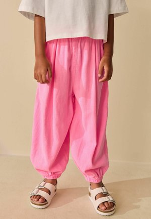 Barn iført løse rosa bukser med elastiske manchetter, hvide sandaler med spænder og en kort hvid skjorte mod en beige baggrund.
