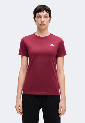 Persoon met kort donker haar die een bordeauxrode The North Face T-shirt en een zwarte broek draagt, staand tegen een effen lichte achtergrond.