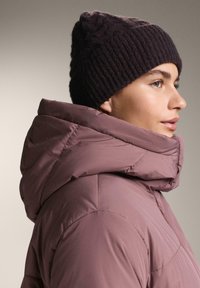 Halvány mályva színű steppelt kabát, magas gallérral, bordázott sötétlila kötött sapkával párosítva. Sima textúrájú és geometrikus varrással díszített.