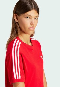 Rød bomulds T-shirt med hvide tre-stribe accenter på skuldrene og et lille logo på brystet. Kortærmet og med rund hals.
