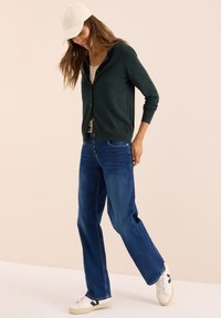 Frau mit weißer Mütze, dunkelgrüner Knopfweste, beigem Oberteil, blauen Jeans und weißen Sneakers vor einem schlichten beigen Hintergrund.