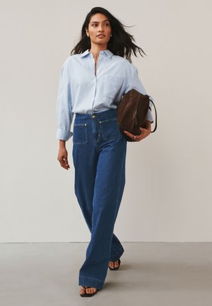 Frau geht, trägt ein hellblau gestreiftes Hemd, hoch taillierte, weit geschnittene Jeans, schwarze Sandalen und hält eine braune Wildledertasche.