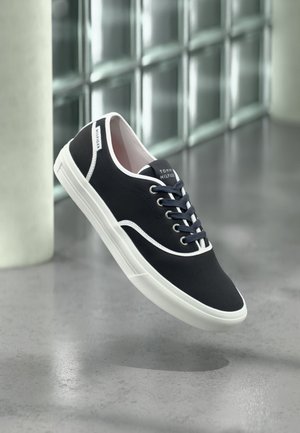 Czarny niski sneaker z białą podeszwą i obramowaniem, sznurowany, z logo "Tommy Hilfiger", unoszący się nad gładką, szarą podłogą i blokami z szkła.