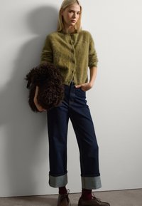 Cárdigan de mohair verde con botones, vaqueros de pierna ancha azul oscuro con los puños enrollados y bolso de piel de pelo marrón. Zapatos marrones y calcetines rojos completan el conjunto.
