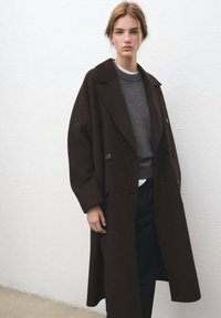 Donkerbruin oversized jas met reverskraag en dubbele rij knopen, gedragen over een grijze trui en zwarte broek. Gladde textuur.