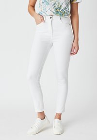 Jeans skinny blancos de cintura alta con cierre de botón, cinco bolsillos y una textura suave. Combinados con zapatillas blancas.