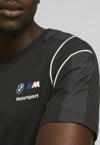 Černé tričko s krátkým rukávem a bílými akcenty, s logem BMW Motorsport na přední straně. Materiál vypadá hladce a má přiléhavý střih.
