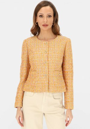 Kobieta w dopasowanej, długorękawowej żółto-pomarańczowej kurtce z tweedu z przodu zapinanej na guziki i kremowych spodniach na jednolitym tle.