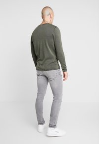 Suéter de manga larga en verde oliva con escote redondo y detalles de costura, combinado con jeans de corte slim en gris claro y zapatillas blancas.