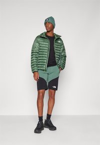 Doudoune verte avec un devant dézippé, T-shirt noir, shorts verts avec des accents noirs et chaussures de randonnée noires. Bonnet en tweed avec logo.