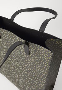 Tote bag med et mørkegrønt og svart bladmønster. Har robuste, svarte håndtak og preget merkevare på fronten. Teksturert materiale.