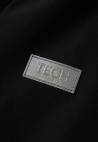 Sort stof med en grå, struktureret etiket prentet med "TECH" og sætningen "name it", omridset med hvid søm.