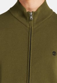 Sudadera de color verde oliva con cremallera, con un cuello y puños acanalados, que presenta un detalle de logo negro en el lado izquierdo. Textura de punto suave.