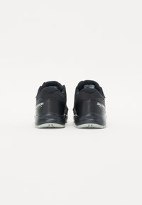 Sapatos atléticos pretos com um tecido de lona texturizado e material sintético, vista traseira mostrando o logotipo e um design minimalista, solas de cor clara.
