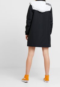 Person som bär en svart och vit huva jacka, står med ryggen synlig, i kombination med orange höga sneakers.