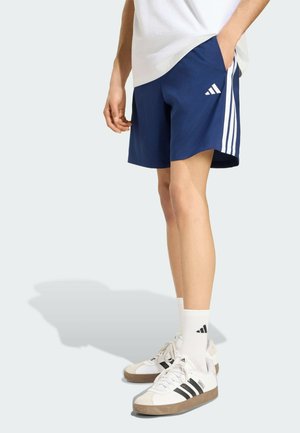 Persona che indossa pantaloncini Adidas blu con strisce bianche, calzini Adidas bianchi e scarpe da ginnastica bianche con strisce nere, in piedi con una mano in tasca.