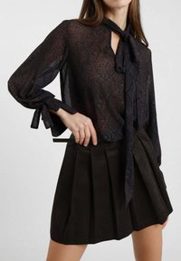 Blusa nera trasparente con stampa serpente, con legacci al collo e alle maniche. Abbinata a una gonna plissettata in suede marrone scuro.