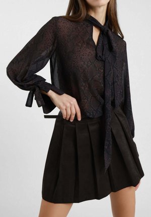 Blusa nera trasparente con stampa serpente, con legacci al collo e alle maniche. Abbinata a una gonna plissettata in suede marrone scuro.