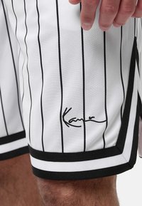 Vita atletiska shorts i vitt med svarta randningar, med en svart fåll och en broderad signatur på den nedre sidan. Ventilerande meshmaterial.