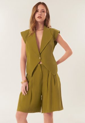 Femme portant un blazer sans manches vert olive avec un bouton et un short taille haute à jambes larges assorti, posant devant un fond uni.