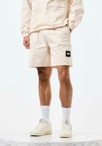 Beige Cargo-Shorts mit seitlichen Taschen und einem schwarzen Markenpatch, kombiniert mit weißen Socken und kremfarbigen Sneakers, auf einer Marmoroberfläche.