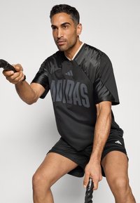 Czarna sportowa koszulka z siateczkowymi wstawkami, z logo "ADIDAS" w ciemnoszarym kolorze. Model nosi ją razem z czarnymi szortami i trzyma liny do ćwiczeń.