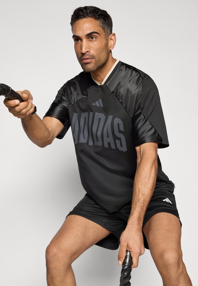 Svart atletisk tröja med meshpartier, med "ADIDAS"-logotyp i mörkgrått. Modellen bär svarta shorts och håller i träningsrep.