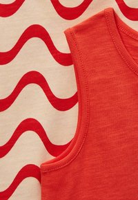 Débardeur en coton rouge avec encolure côtelée, posé sur un tissu crème présentant des motifs ondulés rouges. Les textures contrastent de manière marquée.