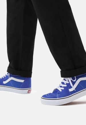 Persoon die zwarte broek en blauwe hoge sneakers met witte veters en zijstrepen draagt en op een witte ondergrond loopt.