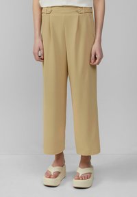 Beige hoge taille broeken met voorplooien en twee knoopdetails. Glad materiaal en wijd uitlopende pasvorm, gecombineerd met platform sandalen.