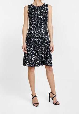 Mouwloze navy jurk met wit bloemmotief, uitlopende rok, gedragen met zwarte, open sandaal met hoge hakken door een staand persoon.