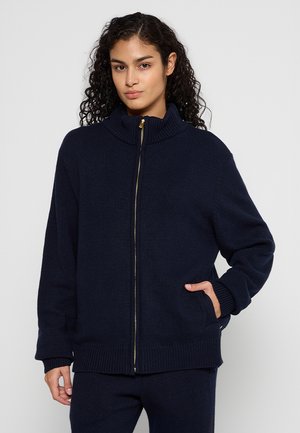 Marine blauwe gebreide zip-up trui met een hoge kraag, geribde manchetten en zoom. Voorzien van een gouden rits en zijzakken. Zacht, gestructureerd materiaal.