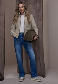 Grauer Wollmischungs-Blazer mit zwei Fronttaschen, weißes Oberteil, blaue Jeans und beige Sneakers, alles vor neutralem Hintergrund.