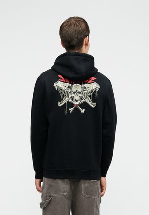 Μαύρη φόρμα με κουκούλα που διαθέτει γραφικό κράνους και σταυροκόκκαλου στην πλάτη, με κόκκινες λεπτομέρειες. Το υλικό φαίνεται απαλό; ο σχεδιασμός είναι καθημερινός και στενός.