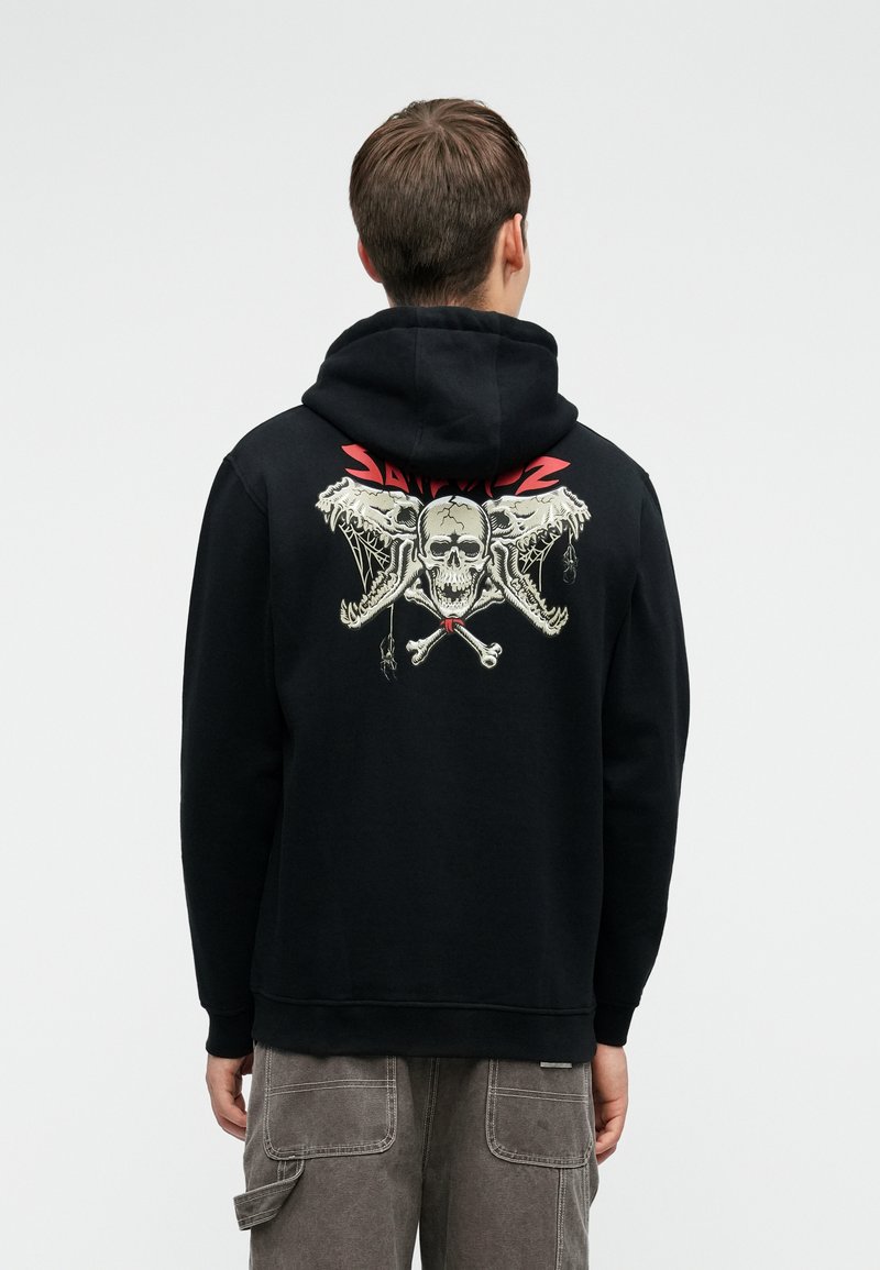 Crna kapuljača s grafikom lubanje i ukrštenih kostiju na leđima, s crvenim naglascima. Materijal izgleda mekano; dizajn je ležeran i prilagođen tijelu.