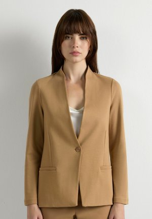 Frau mit braunen Haaren trägt einen beigefarbenen Blazer mit einem Knopf über einem weißen Oberteil und steht vor einem schlichten, hellgrauen Hintergrund.