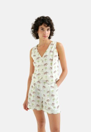 Mujer vistiendo un conjunto de chaleco sin mangas blanco abotonado y pantalones cortos con un pequeño estampado de elefante y palmera, de pie con una mano en el bolsillo.