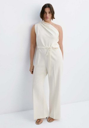 Femme portant une combinaison écrue asymétrique avec une ceinture à la taille, jambes larges et sandales, debout devant un fond blanc uni.