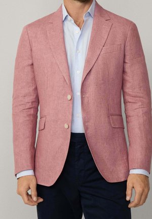 Mand iført en lyseblå skjorte med knapper, lyserød blazer med to knapper og mørke bukser, stående mod en neutral baggrund.