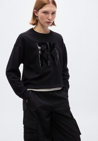 Μαύρο φούτερ με ανάγλυφο λογότυπο DKNY από παγιέτες, με στρογγυλή λαιμόκοψη και ριμπ τελείωμα. Φορεμένο με μαύρο παντελόνι cargo.