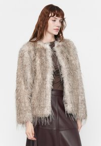 Faux fur jacket in light beige with a textured, fluffy design, cropped sleeves, and an open front, paired with a fitted brown top and burgundy skirt.