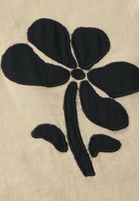 Applicazione floreale in tessuto nero su sfondo beige, caratterizzata da cinque petali e un cerchio centrale, con bordi texturizzati.