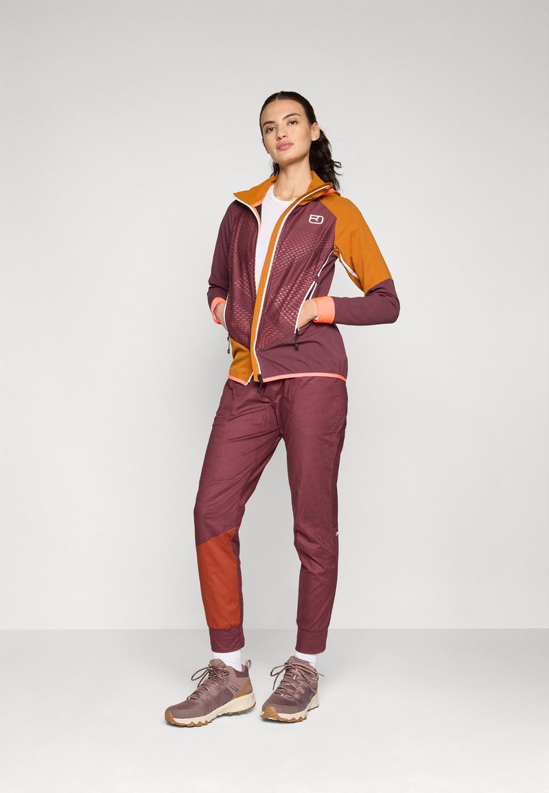 Sportliches Outfit mit einer lila-orangefarbenen Jacke mit Reißverschluss und Netzstruktur, ergänzt durch passende Jogginghosen und graue Wanderschuhe.