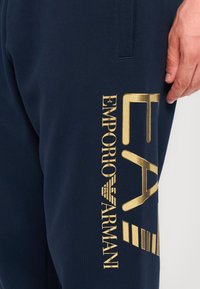 Navy sweatpants med et stort guld "EA" logo og "Emporio Armani" tekst. Fremstillet af blødt stof med en glat tekstur, detaljeret med striber.