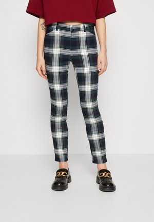 Personne portant un pantalon à carreaux noirs, blancs et verts, des mocassins noirs avec des chaînes dorées, et un haut rouge court, debout devant un fond uni.